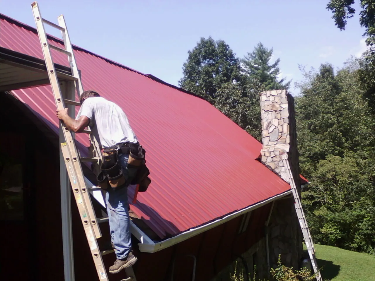 Roofer on a ladder beside a red metal roof and stone chimney — Soffit & Fascia Repair by Roofing Brothers in Totowa